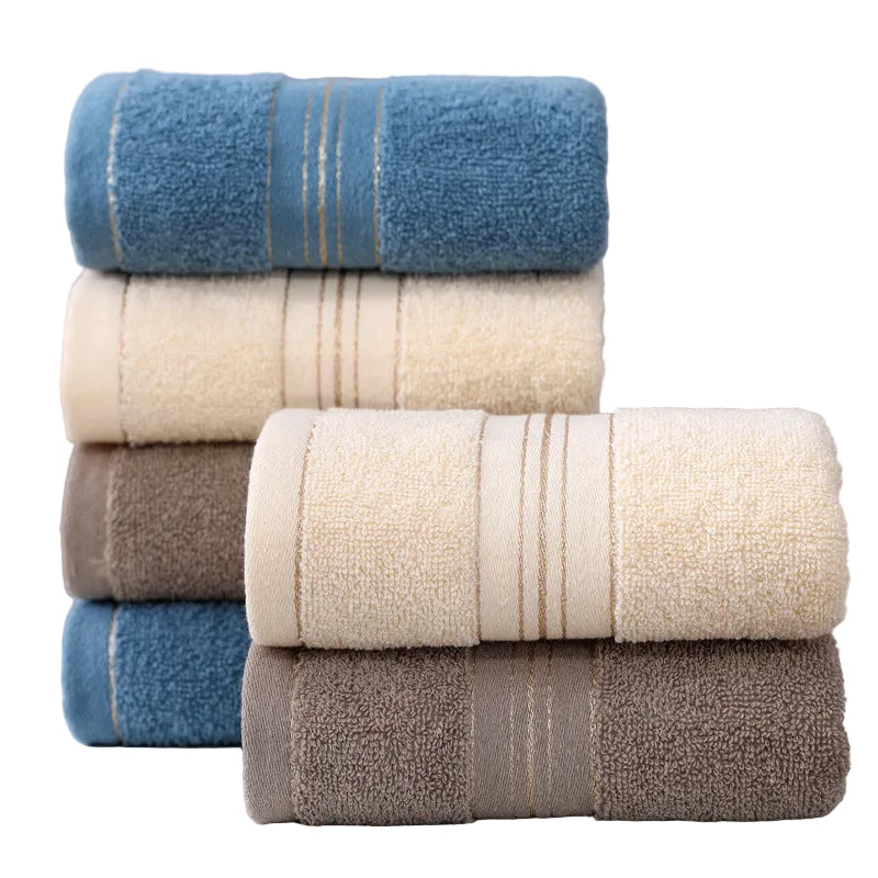 Four stacks of folded cotton towels in blue, cream, taupe, and brown, each with a central striped band, displayed on a light surface against a soft gray background.