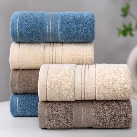Four stacks of folded cotton towels in blue, cream, taupe, and brown, each with a central striped band, displayed on a light surface against a soft gray background.