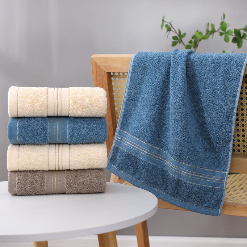 Four folded towels stacked on a white round table next to a wooden chair, with one blue towel draped over the chair. The towels have textured terry cloth fabric and double stripe borders in beige and blue.