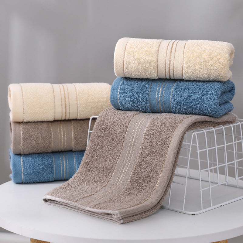 A set of folded and rolled towels in neutral tones, including cream, blue, taupe, and brown, arranged on a white table with a wire stand against a soft gray background.