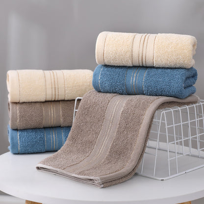 A set of folded and rolled towels in neutral tones, including cream, blue, taupe, and brown, arranged on a white table with a wire stand against a soft gray background.