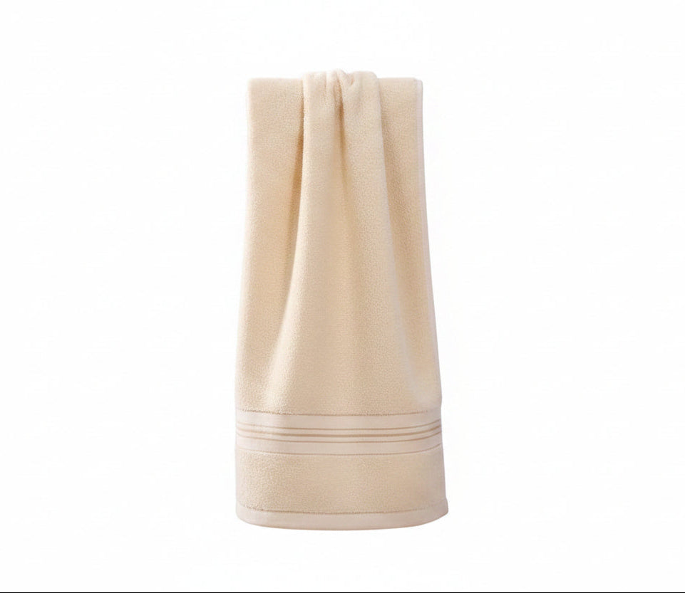 A beige cotton bath towel with a stitched border sits draped over a wooden chair.