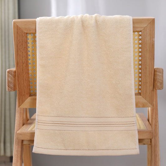 A beige cotton bath towel with a stitched border sits draped over a wooden chair.